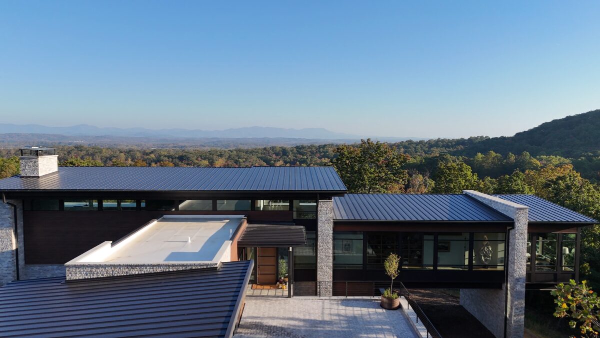 A modern, flat-roofed luxury home with sleek metal roofing and large glass windows, set against a backdrop of lush green forest and distant mountain ranges under a clear blue sky. The home features stone and wood accents, with a spacious, paved entryway and potted plants adding a touch of greenery to the contemporary design.