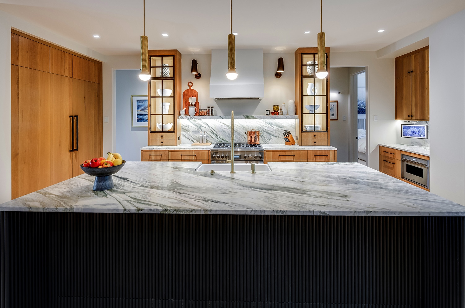 Elegant luxury kitchen in a custom home in Greenville, SC, designed and built by Bluestone Construction. The centerpiece is a Calacatta Manhattan marble island with a fluted black cabinetry base, creating a striking contrast. Warm white oak cabinetry and glass-front display shelving add sophistication, while modern brass pendant lights enhance the ambiance. A professional-grade Thermador gas range is framed by a quartzite backsplash and custom range hood, seamlessly blending luxury and functionality. Thoughtful details include a hidden pantry, built-in microwave, and under-cabinet lighting, showcasing exceptional craftsmanship.